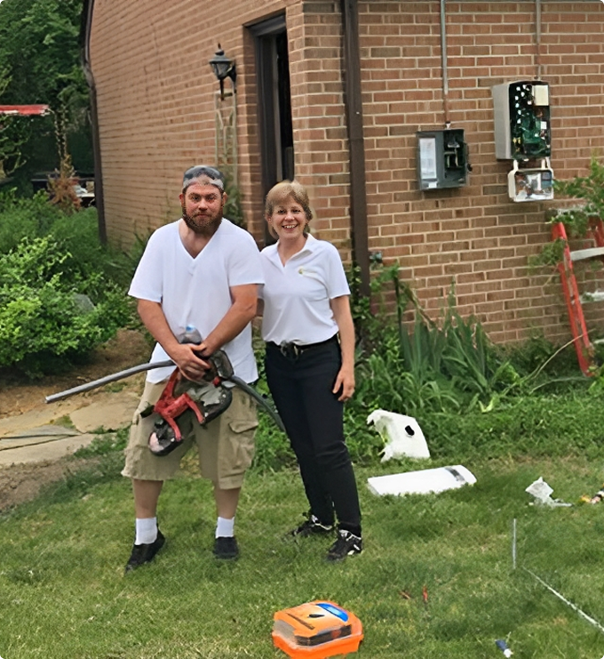 Two people standing outside near a brick building, surrounded by tools and equipment. One person holds a piece of machinery, and both are looking at the camera.