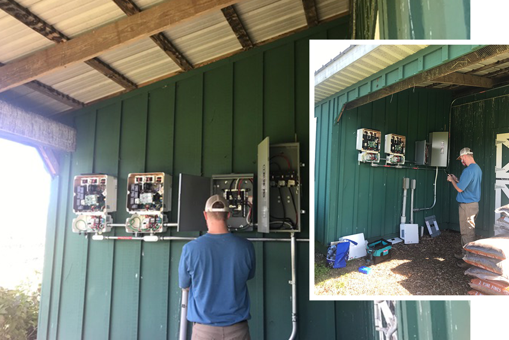 A person in a blue shirt works on electrical equipment mounted on the exterior green wall of a shed; tools and supplies are on the ground nearby.